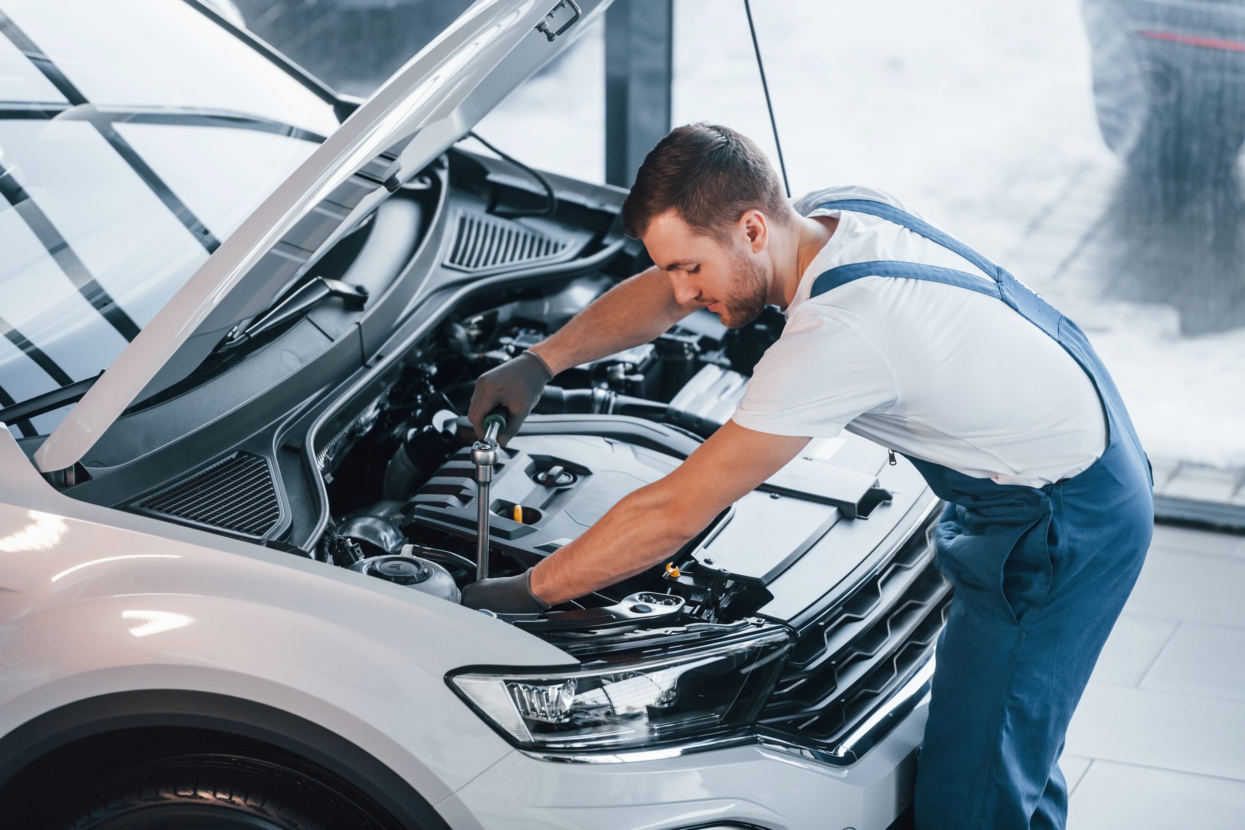 Hood is opened. Young man in white shirt and blue uniform repairs automobile